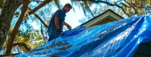 a homeowner covers a damaged roof with a blue tarp to protect their belongings from water damage after a storm